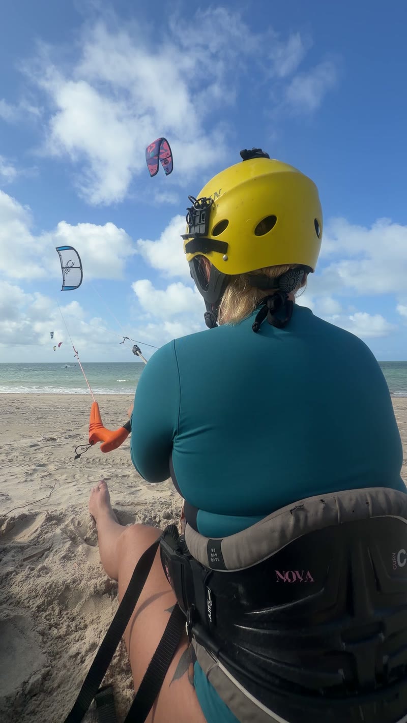 Aluna de capacete amarelo sentada na areia pilotando o kite, com vários kites no céu de Cumbuco