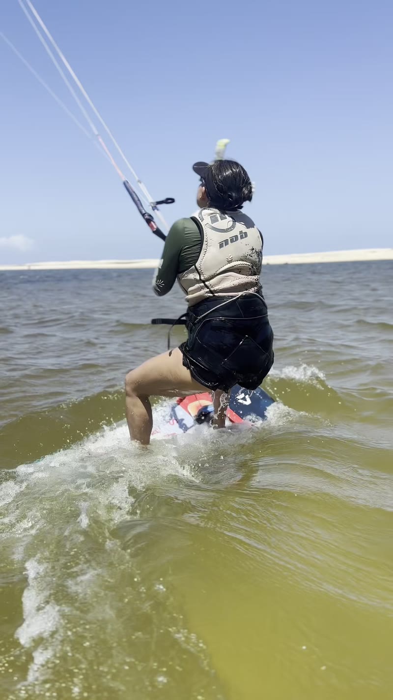 Aluna velejando de kite em Cumbuco, filmada da água, com o kite no céu e a praia ao fundo