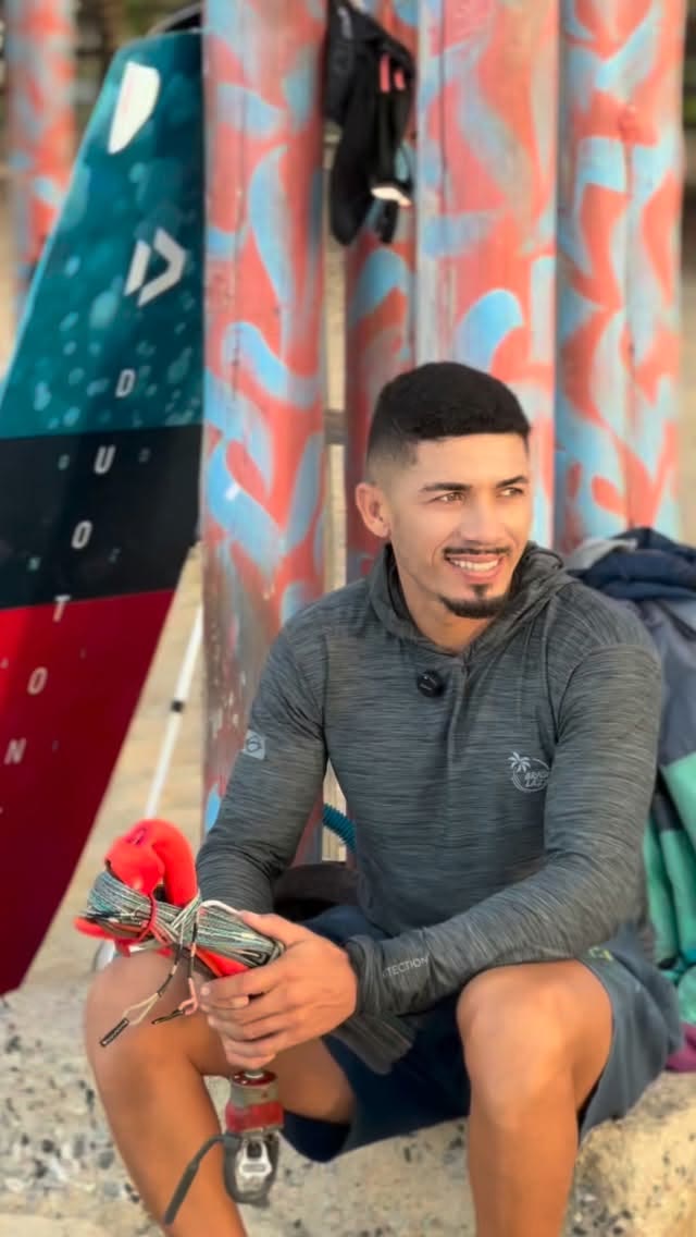 João sentado com as linhas do kite nas mãos, pranchas ao fundo