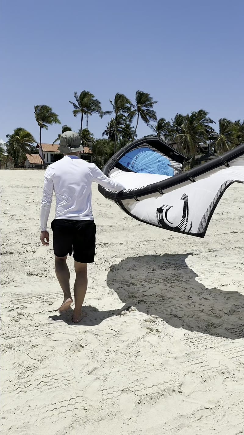 Aluno atravessando a areia de Cumbuco com o kite na mão, coqueiros curvados pelo vento